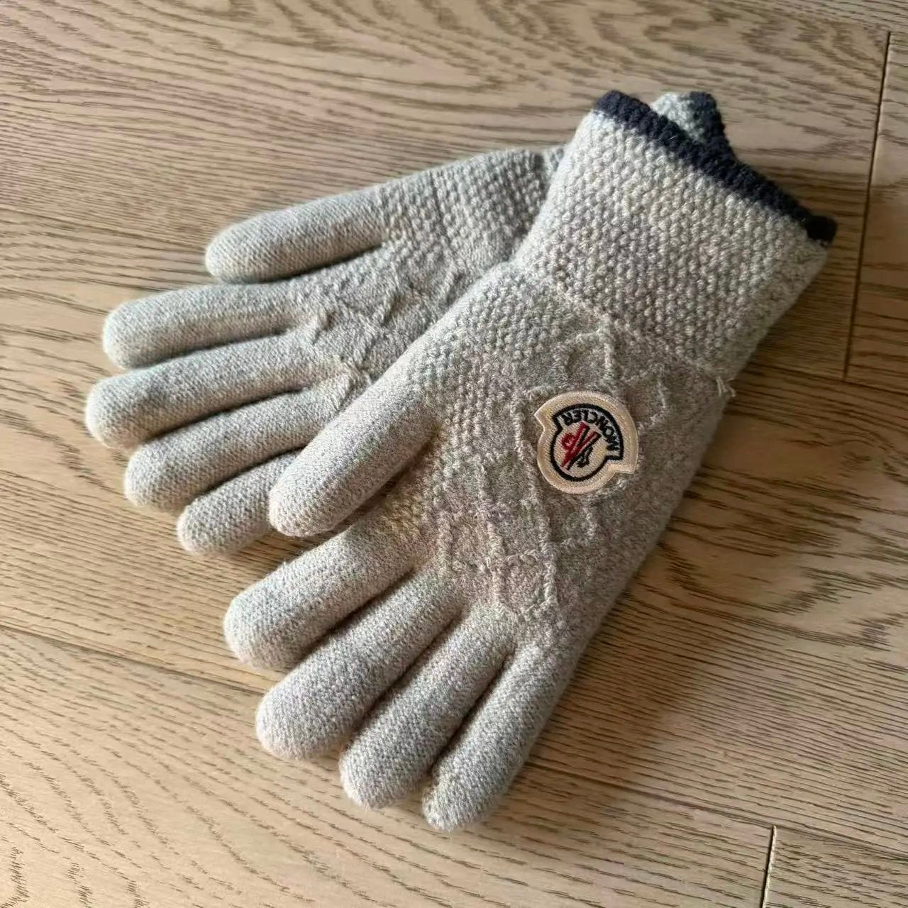 A pair of knit winter gloves in a light beige color with a dark navy cuff, featuring a circular Moncler logo patch on the back of one glove, placed on a light wood floor.