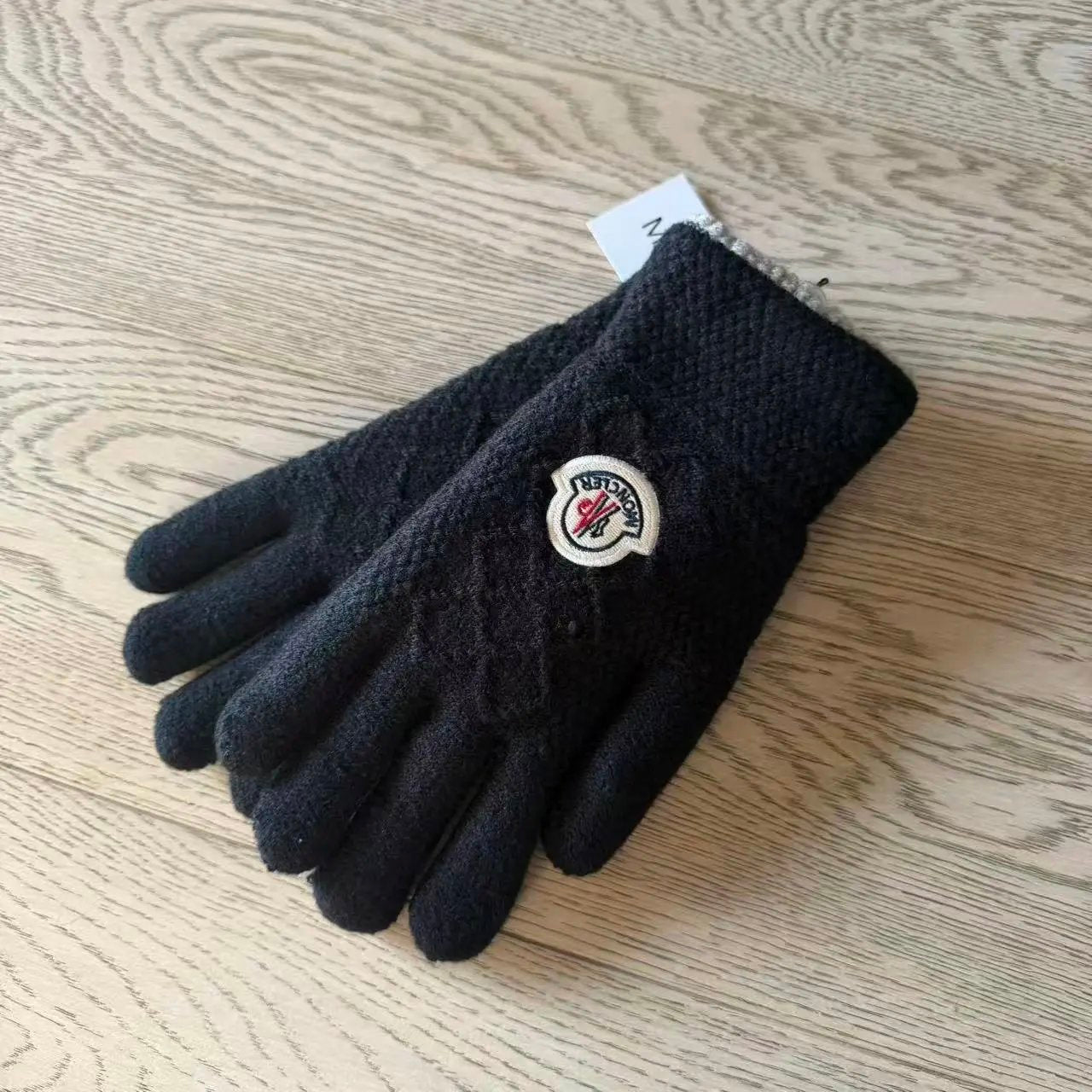 A pair of black knit winter gloves with a cable-knit pattern and a circular Moncler logo patch, displayed on a light wood grain surface.