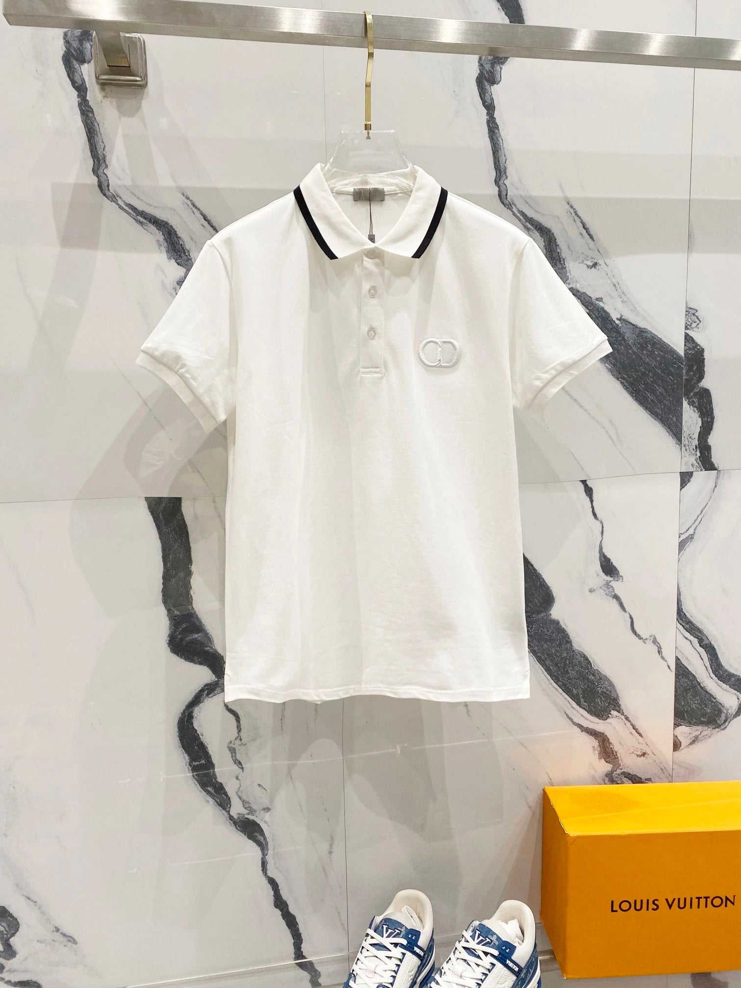 White polo shirt with black trim on a marble wall background, with a Louis Vuitton box in the foreground.