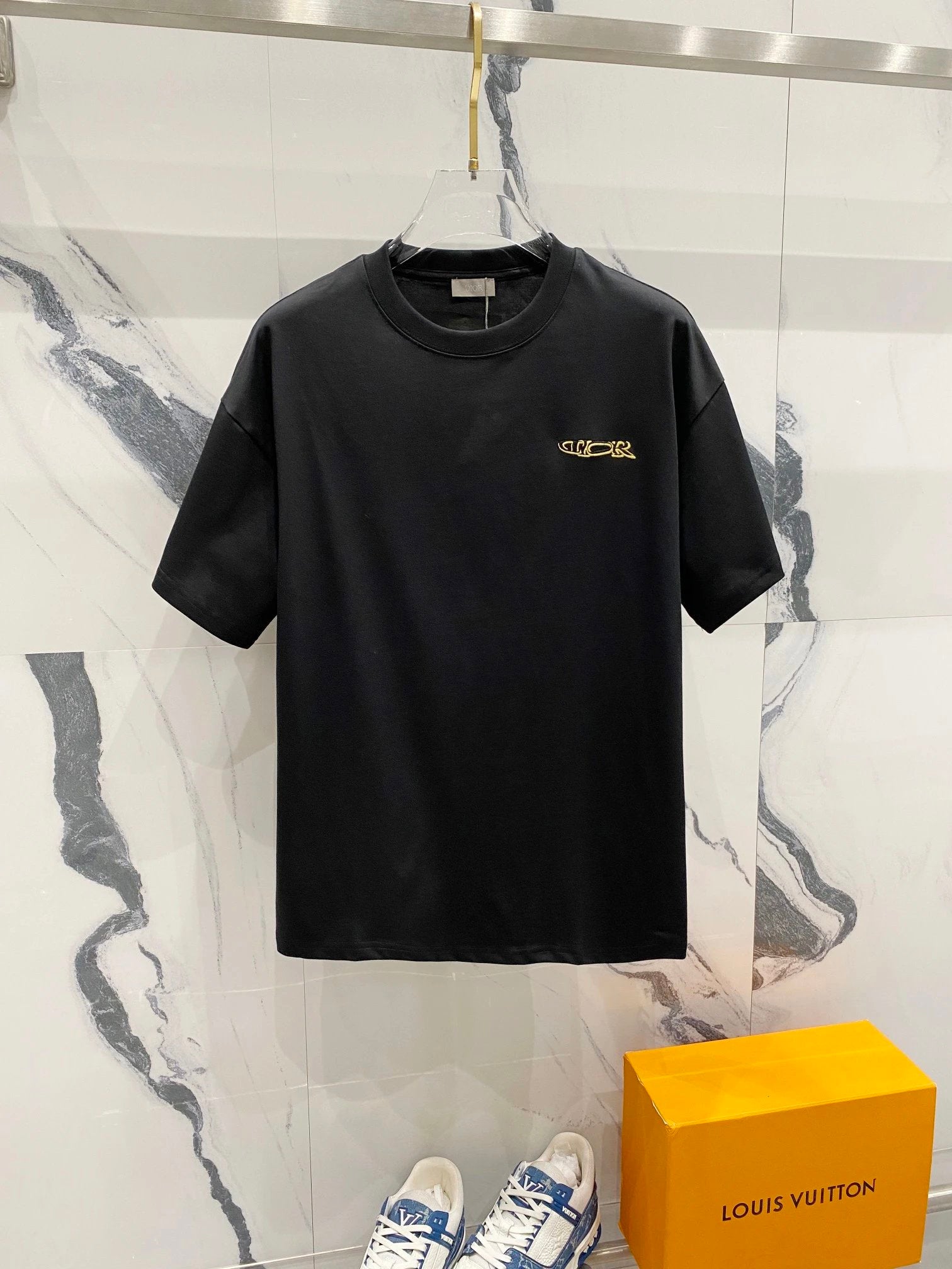 Black t-shirt with a yellow logo on a hanger against a marble wall with a Louis Vuitton box below.