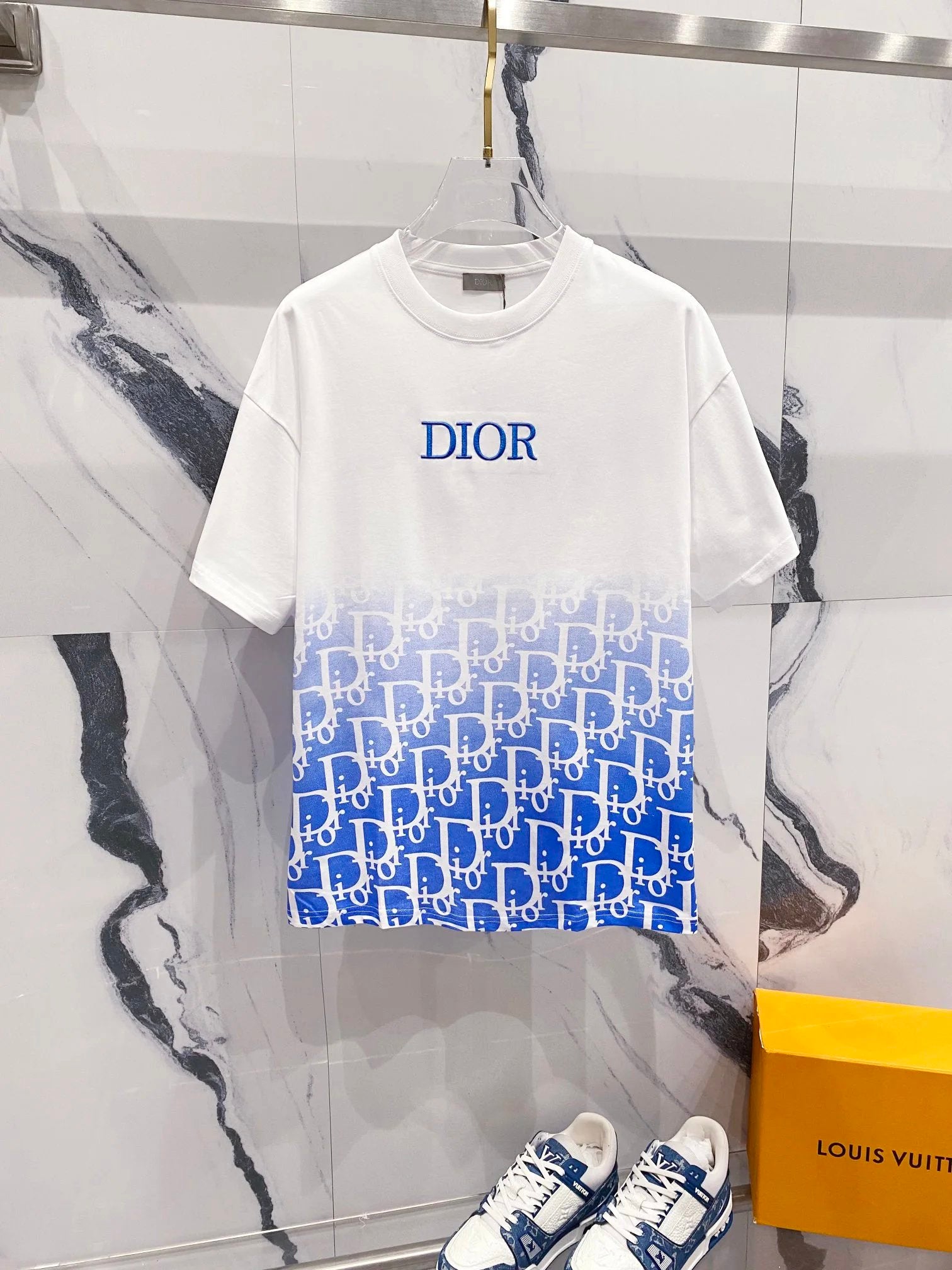 White t-shirt with blue 'Dior' logo and pattern, paired with white sneakers, on a marble surface.
