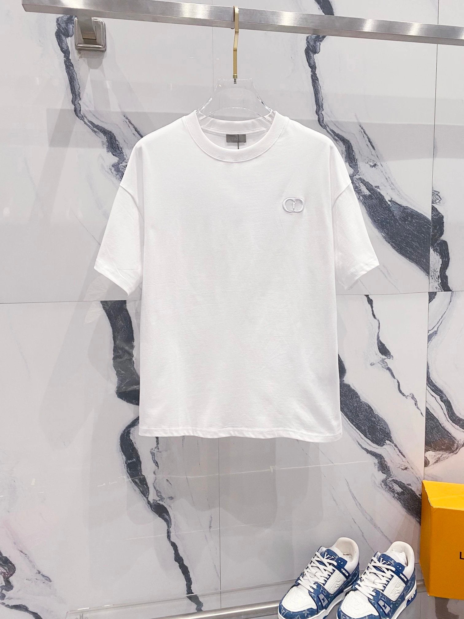 White t-shirt with a small logo on a hanger against a marble wall.