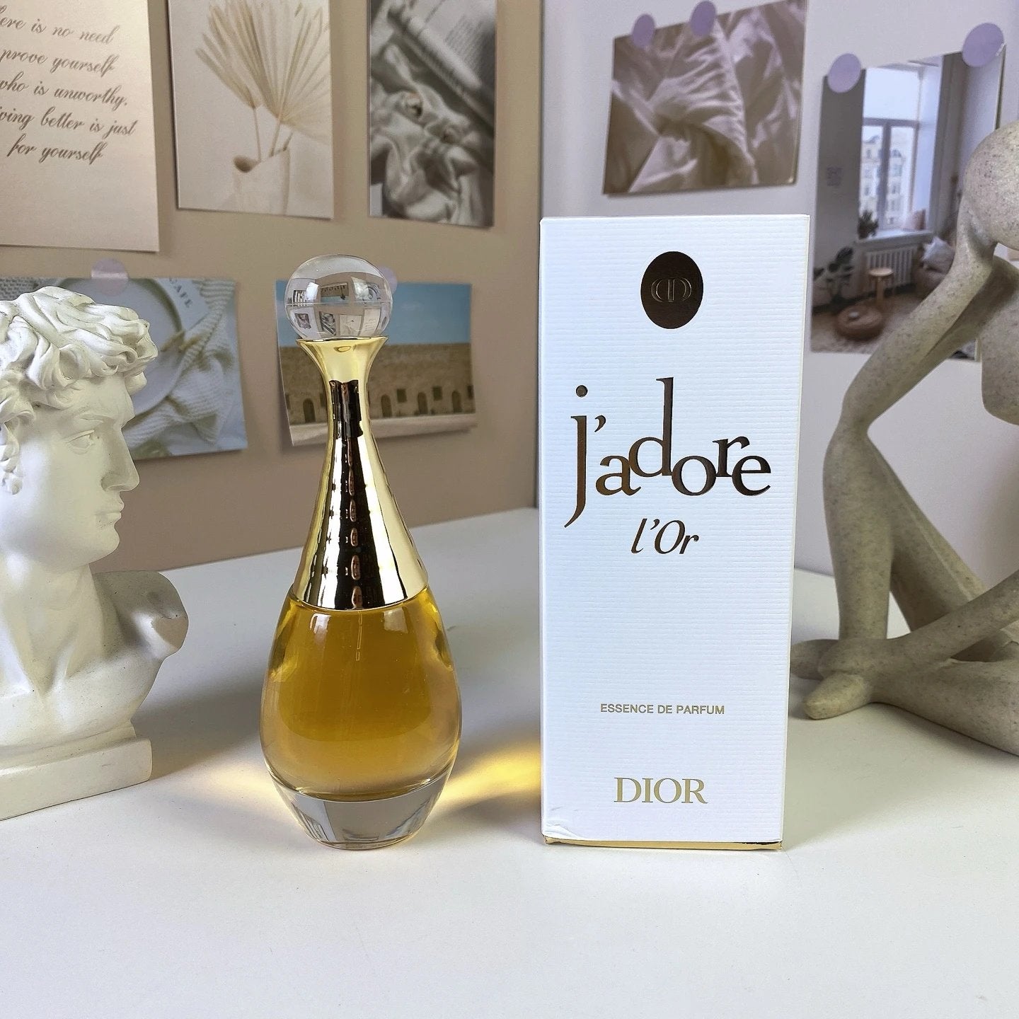 Dior j'adore l'Or perfume bottle and packaging on a decorative surface with sculptures and photos in the background.