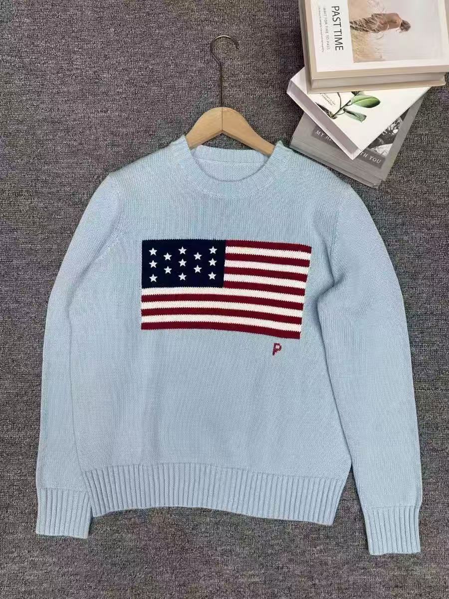 Light blue sweater with American flag design on a hanger against a gray background.