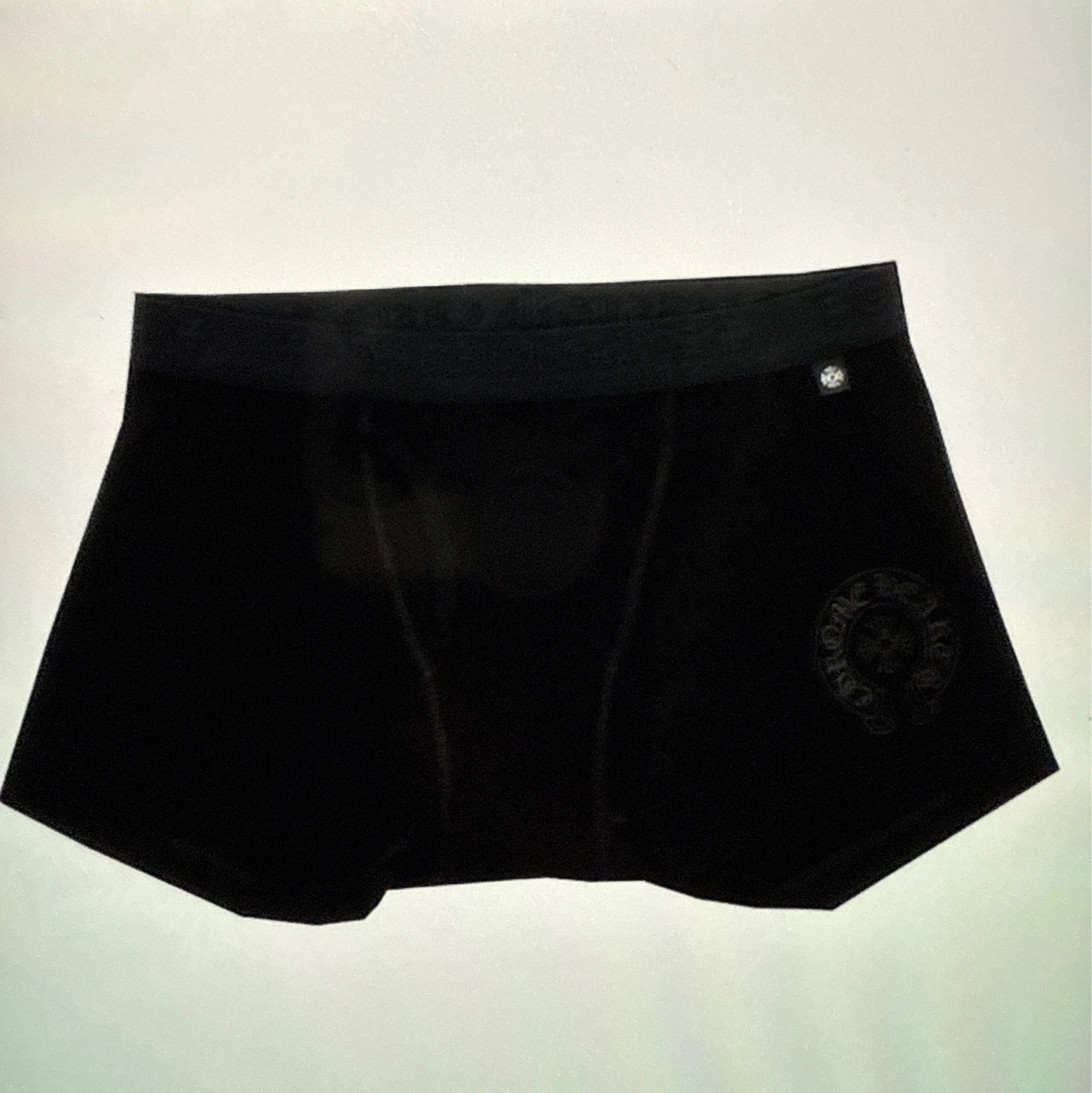 Black boxer briefs with a small silver button on the right hip and a dark circular logo patch on the left leg, shown against a light off-white background.