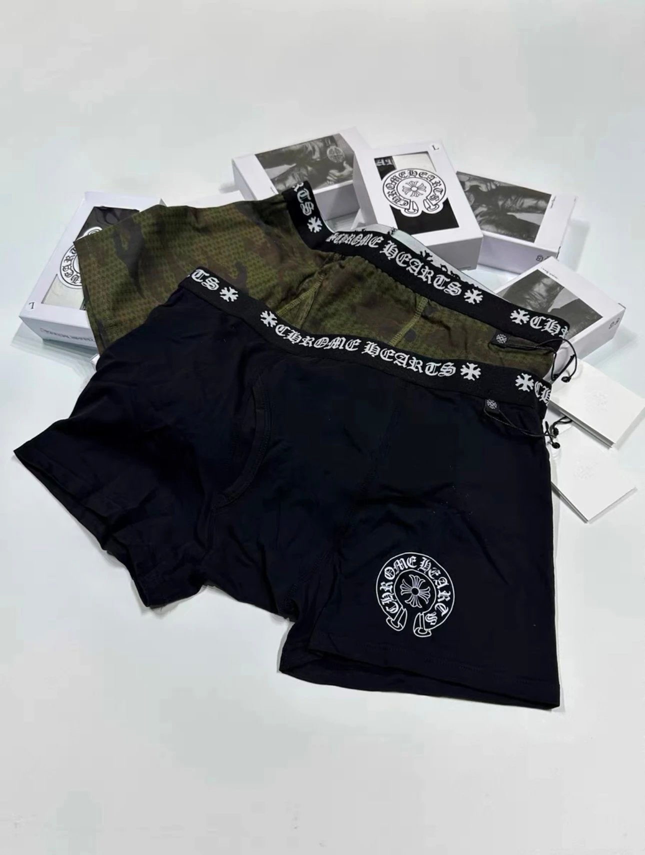 Three pairs of boxy boxer briefs from Chrome Hearts are arranged on a white surface with their branded packaging boxes. The foreground pair is black with a white circular logo, the middle pair is olive green with a black patterned waistband, and the background pair is dark gray with a similar patterned waistband.