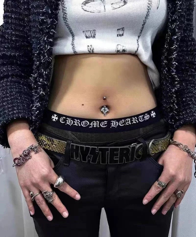 A close-up lifestyle shot of a person's midsection and hands, showcasing a black gothic-style belt with 'CHROME HEARTS' text and a snakeskin-patterned 'HYSTERIC' belt. The person is also wearing a white ribbed crop top, a black-and-white tweed jacket, and multiple silver rings.