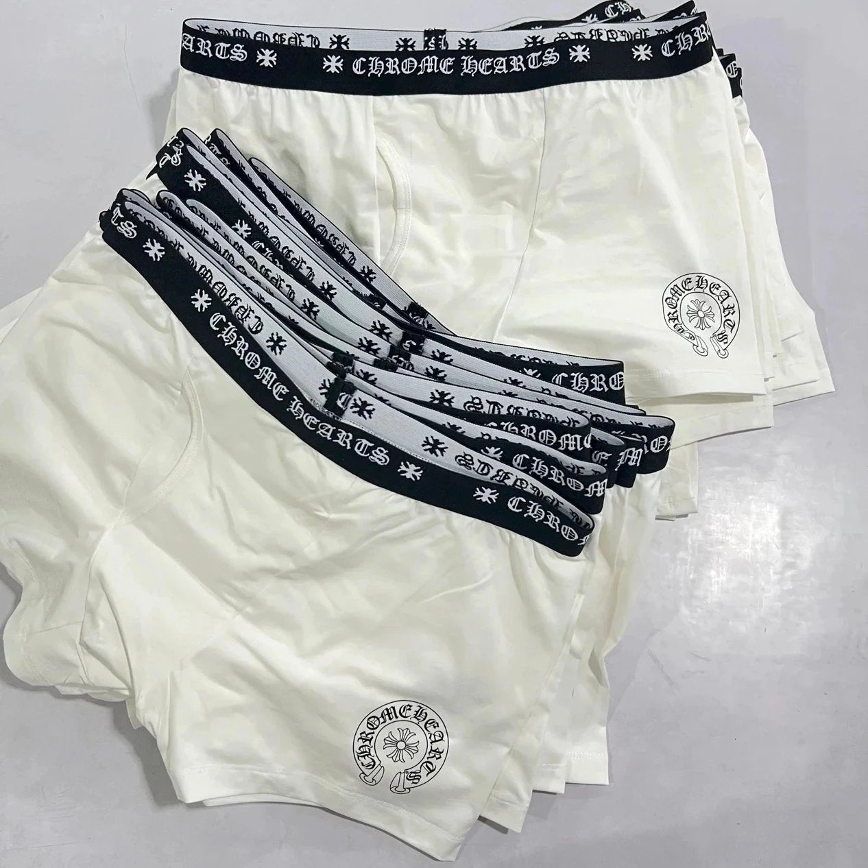 A stack of five men's boxer briefs in off-white, with black elastic waistbands featuring a repeating white logo pattern. The front pair is fanned out to show the leg openings and a circular logo printed near the lower left leg.