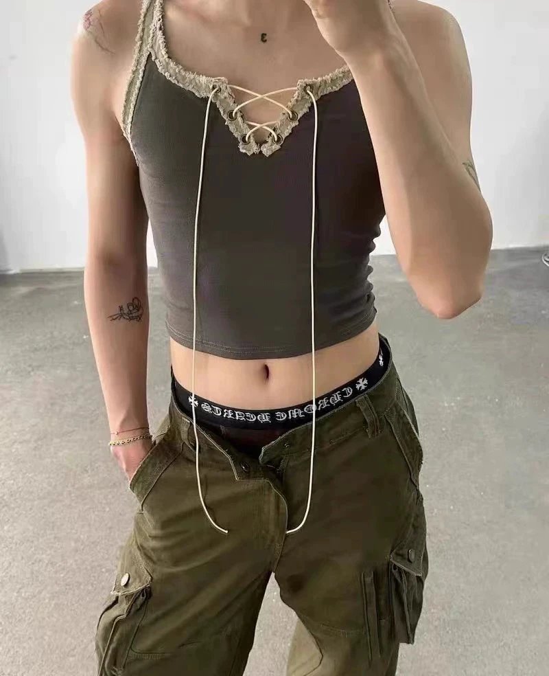 A person wearing a dark olive-brown lace-up crop top and matching cargo pants, photographed indoors against a plain light gray wall and concrete floor.