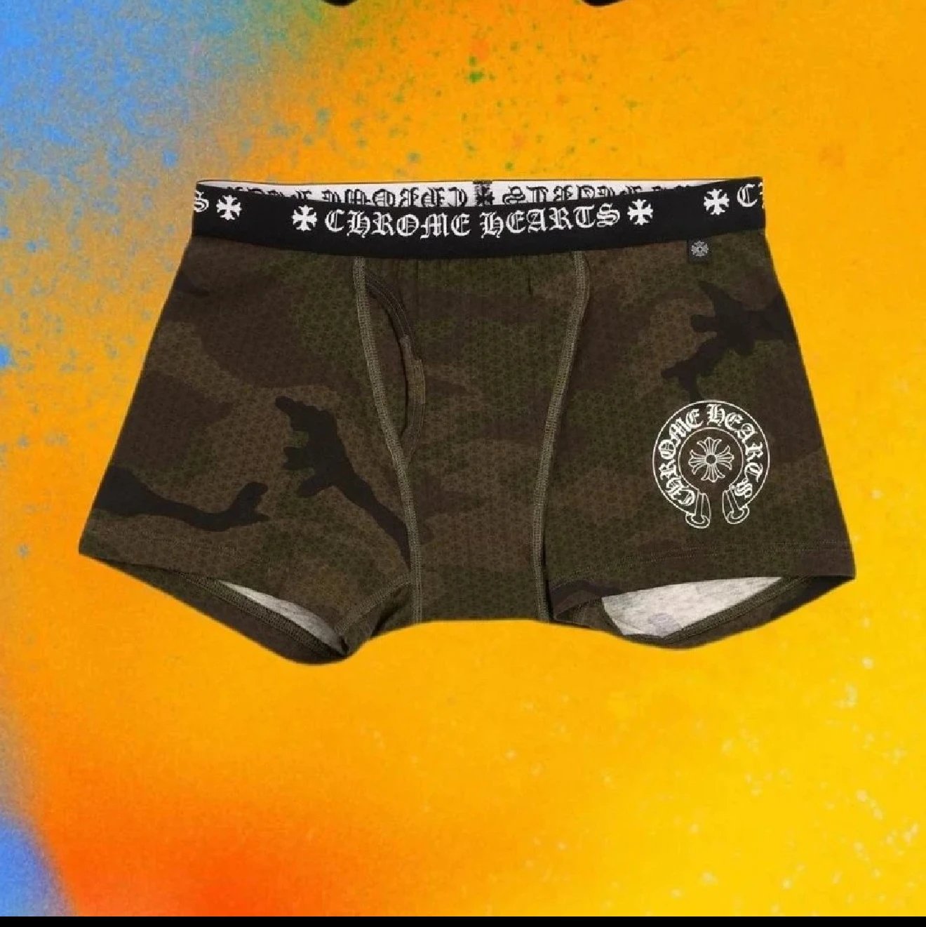 Chrome Hearts men's boxer briefs featuring a dark green and black camouflage pattern, a black waistband with the brand's gothic logo, and a white circular logo patch on the left leg, displayed against a vibrant orange and yellow background.