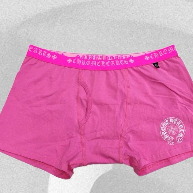 Bright pink boxer briefs with a matching pink elastic waistband printed with the word 'Chrome' in white, plus a white circular logo on the lower right leg, laid flat on a light gray textured surface.