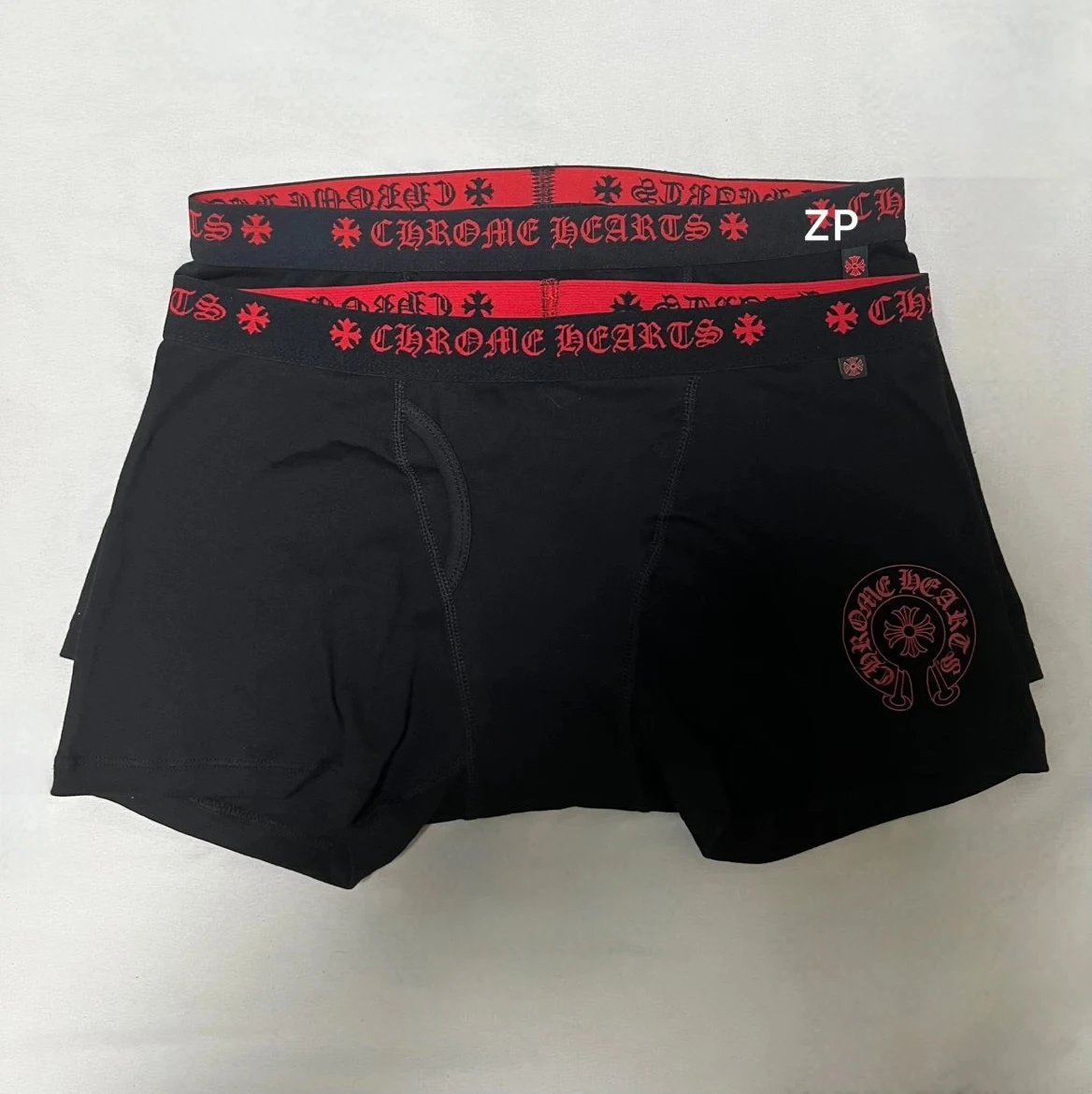 Two pairs of black boxer briefs with red and black patterned elastic waistbands, displayed flat against a light gray background. The waistband features the text 'CHROME HEARTS' and a 'ZP' monogram.