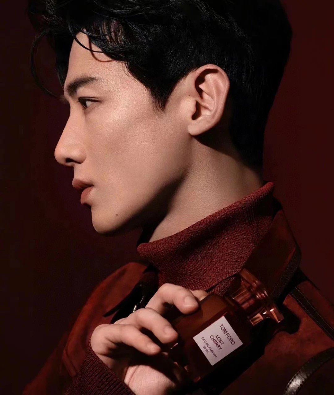 A man in a dark red turtleneck sweater holds a Tom Ford Scorpion perfume bottle against a dark red background.