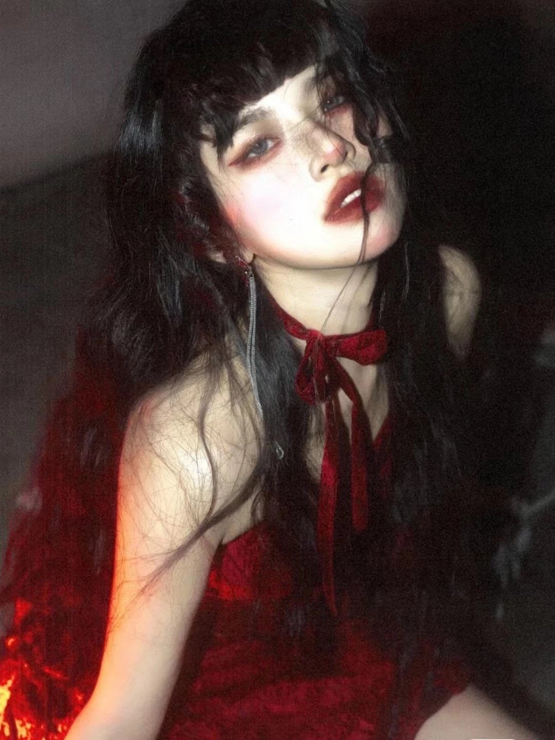 A woman with long dark hair and dramatic red eye makeup wearing a red lace top and red neck tie, captured in moody low-light lighting with a dark background.