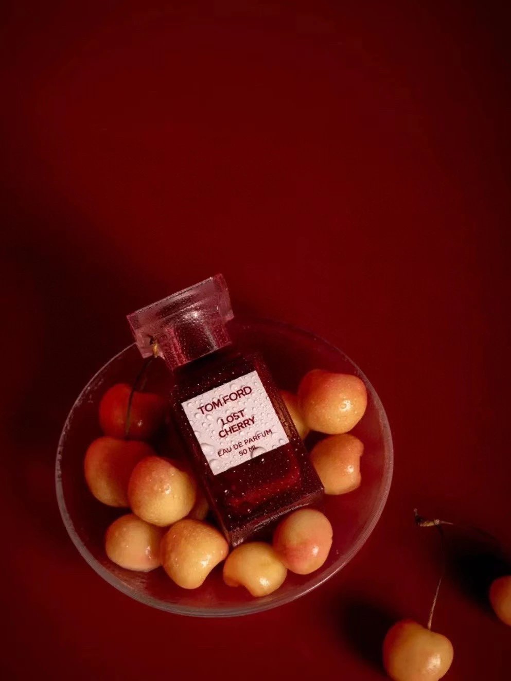 A bottle of Tom Ford Lost Cherry perfume oil with a clear cap, resting on a glass bowl filled with cherries against a dark red background.