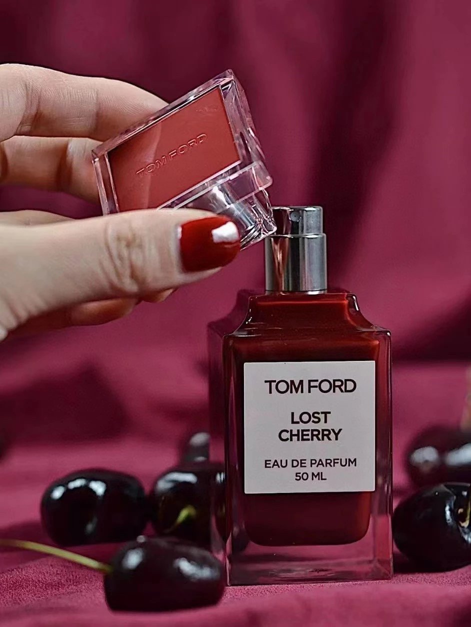 A hand with red nail polish holds an open Tom Ford lipstick compact above a Tom Ford Lost Cherry Eau de Parfum bottle, with dark cherries scattered on a deep burgundy fabric background.