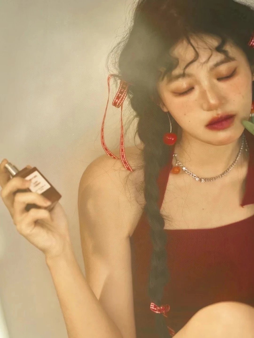 A woman with dark curly hair wearing a red top and red accessories, holding a brown rectangular perfume bottle. The photo has a warm, hazy, vintage aesthetic with soft lighting.