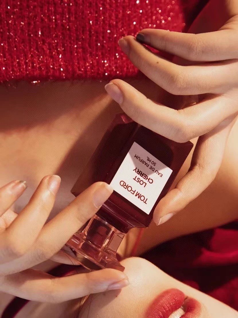 A close-up of a person holding a Tom Ford Lost Cherry Eau de Parfum bottle. The person is wearing a sparkly red top and red lipstick, with their hands and lips clearly visible against a warm, red-toned background.