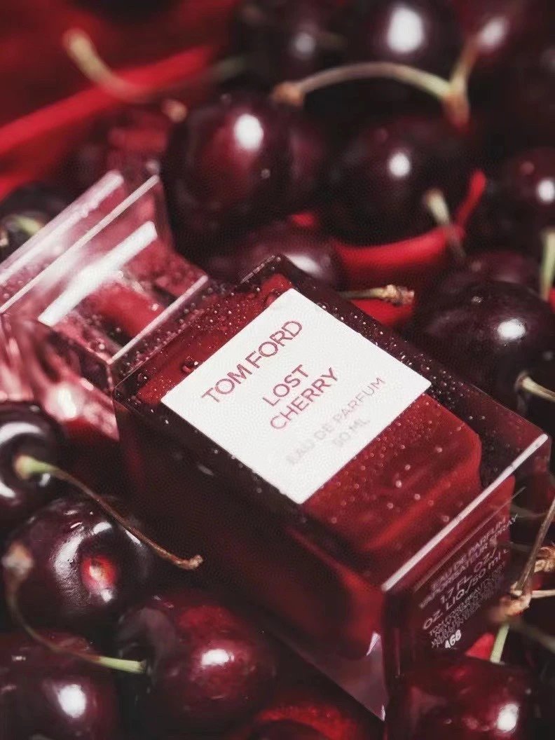 A Tom Ford Lost Cherry Eau de Parfum bottle surrounded by fresh dark red cherries. The clear glass bottle shows deep red liquid inside and features a white label with red text.