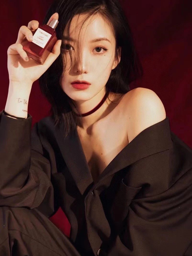 A woman in a dark off-the-shoulder top holds up a small rectangular perfume bottle with a red liquid and a gold cap against a deep red background.